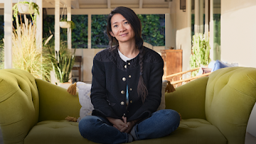 Director, screenwriter, editor, and producer Chloé Zhao, wearing a blue jacket, sitting on a green couch in a backyard on a sunny day.